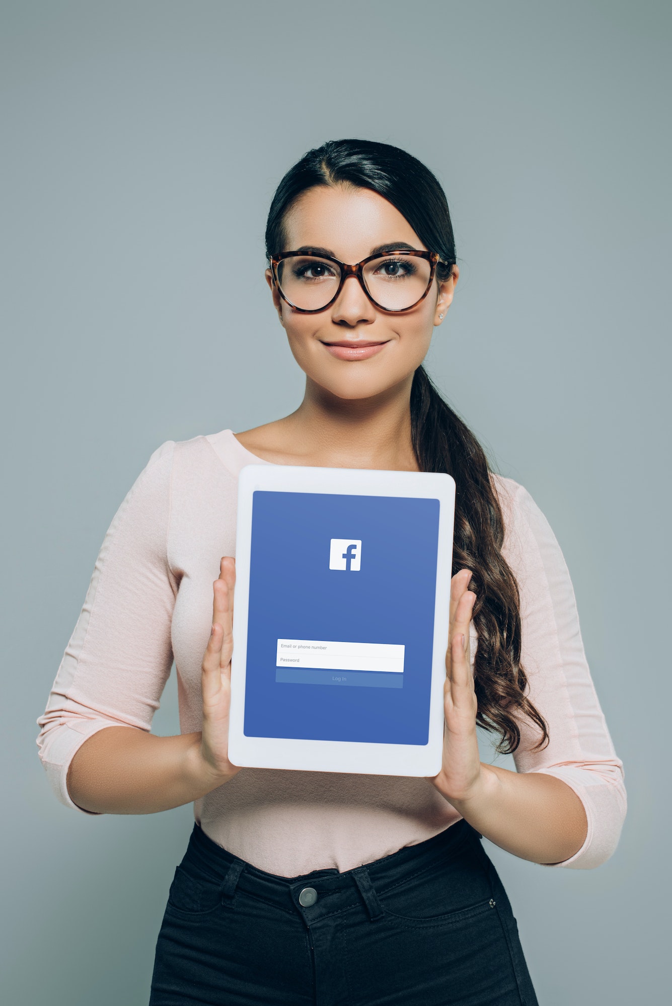 woman in eyeglasses showing digital tablet with facebook appliance, isolated on grey