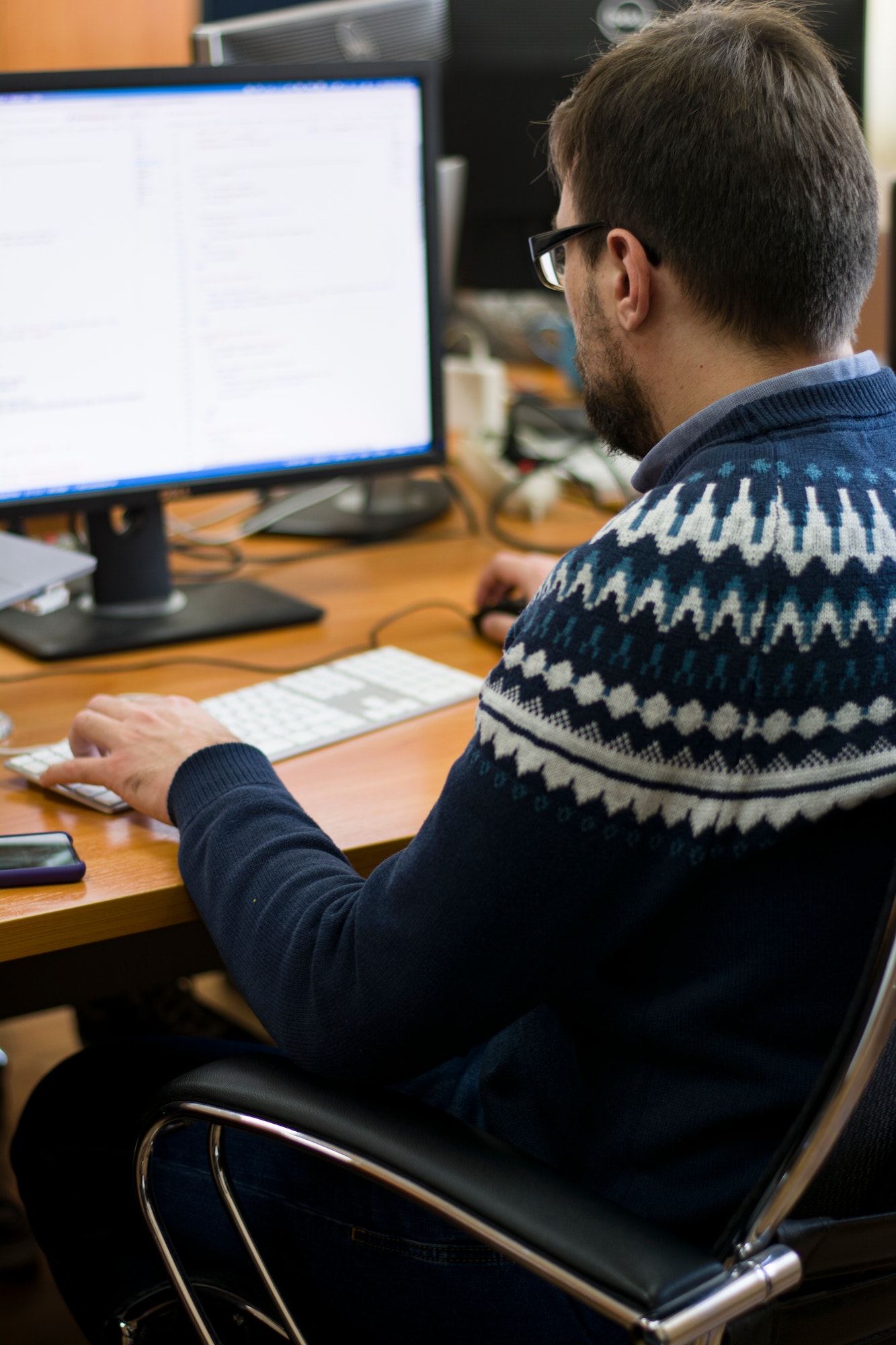 Young man with glasses sits in the office in front of his laptop and monitor working on it