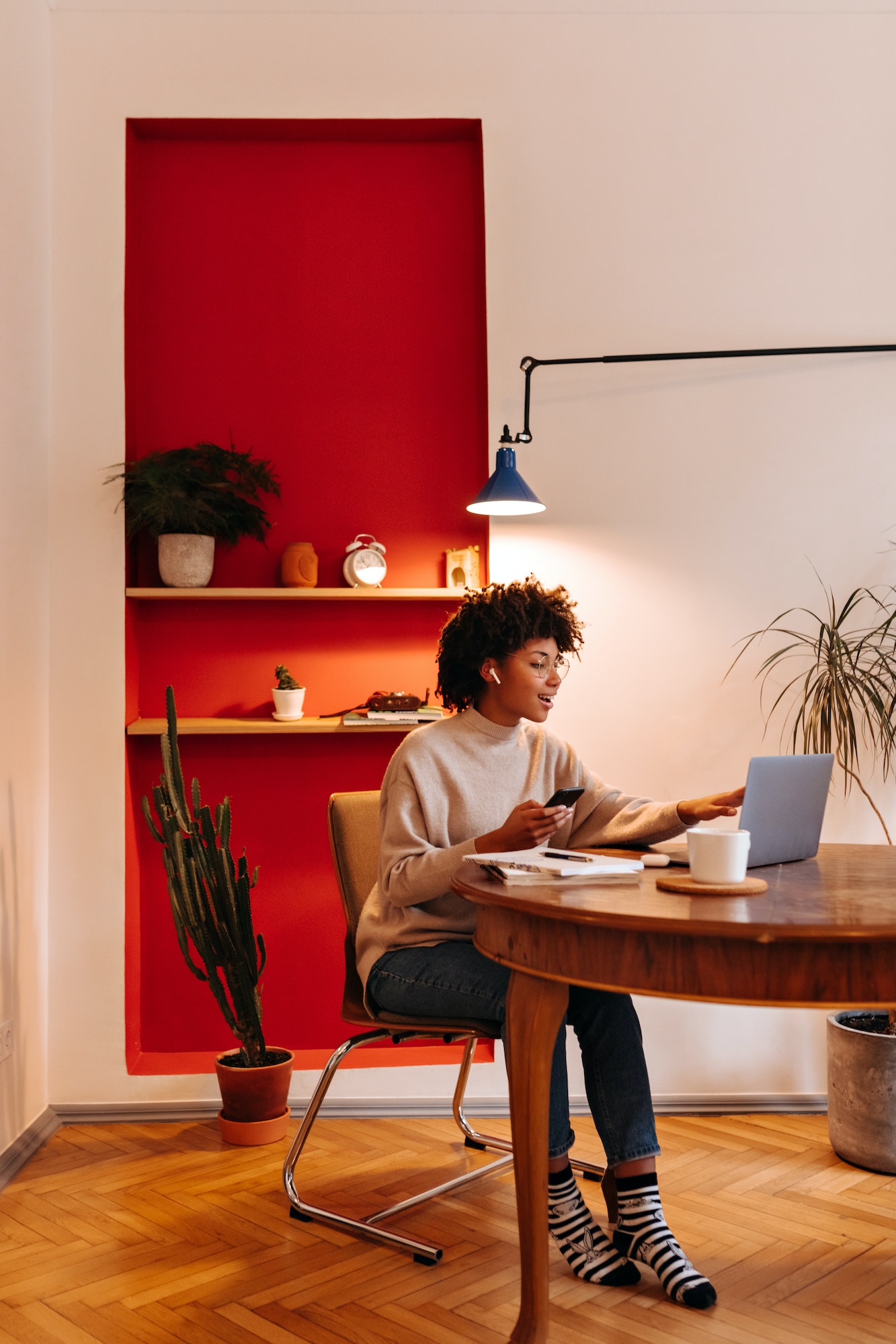 Young woman in sweater communicates via video link in laptop, holds smartphone and sits at table in