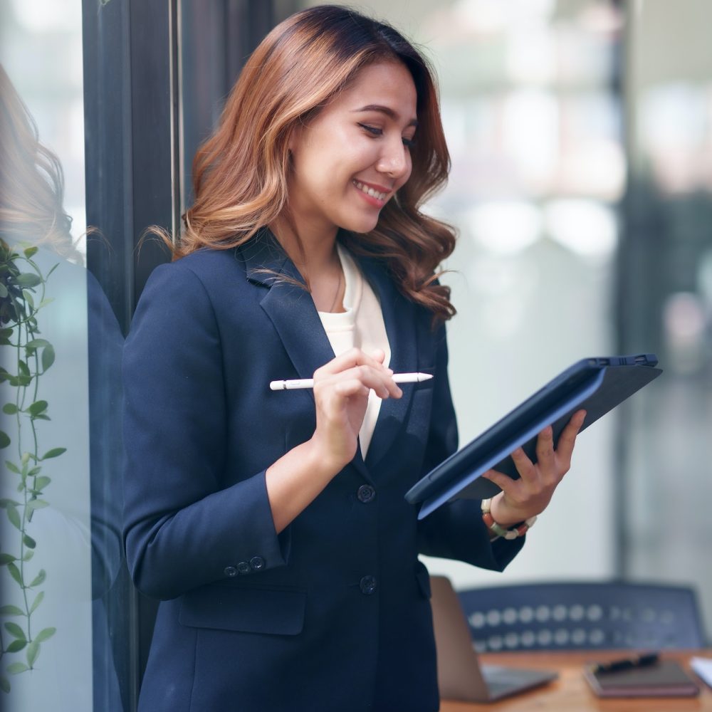 Attractive smiling Asian businesswoman standing holding tablet working and recording work details